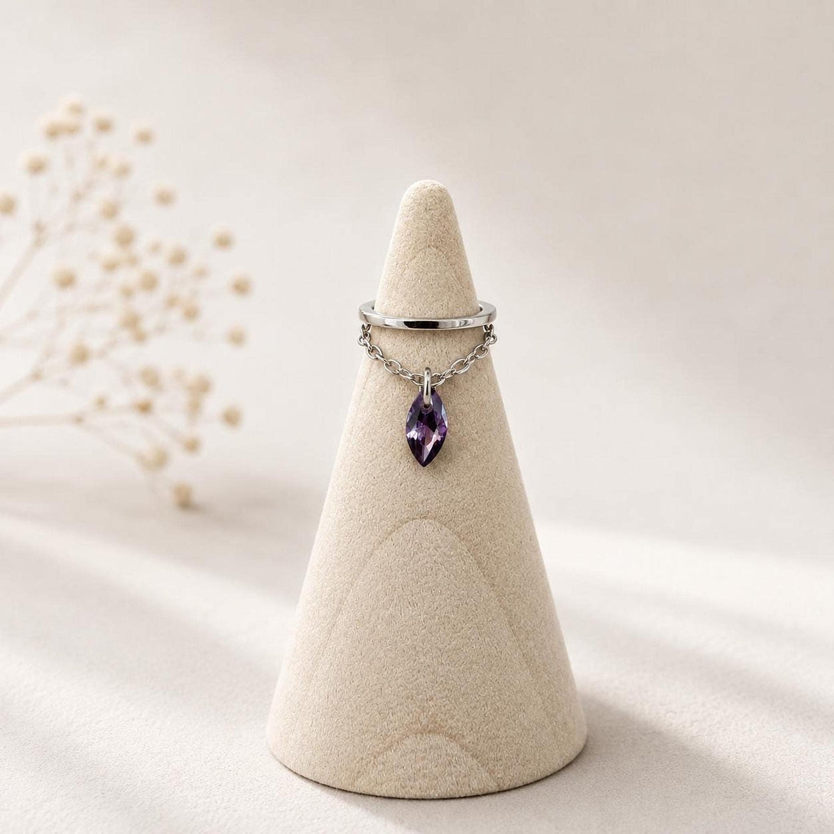 A silver ear cuff with a dangling chain and a purple marquise gemstone displayed on a beige ring cone.