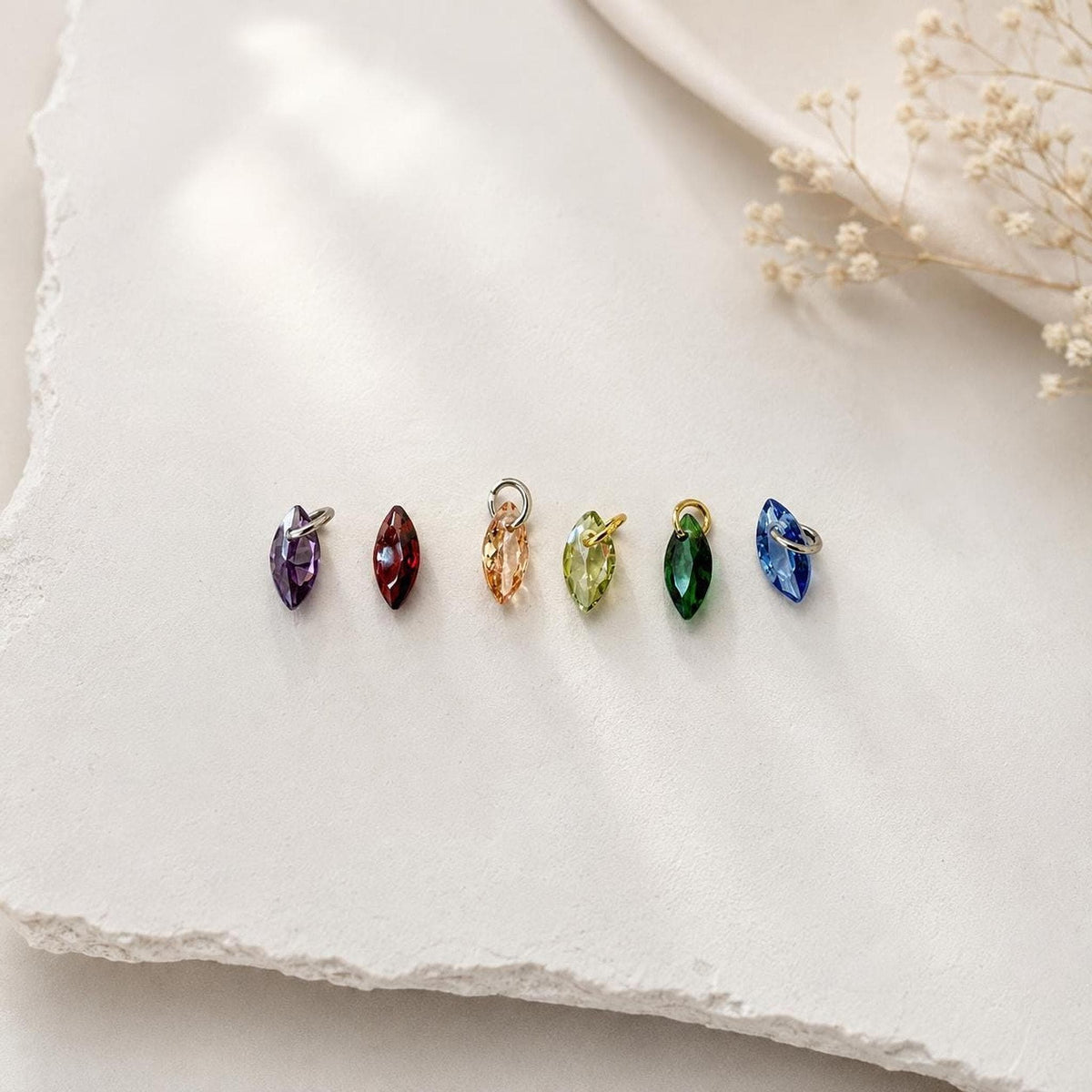 Six marquise-cut gemstone charms in various colors, including purple, red, peach, green, and blue, laid out on a textured white surface.