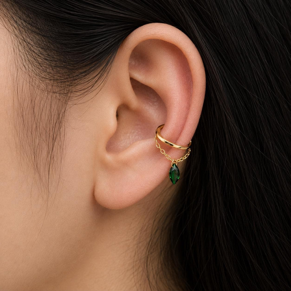 A gold ear cuff with a hanging chain and a green marquise gemstone worn on a human ear.