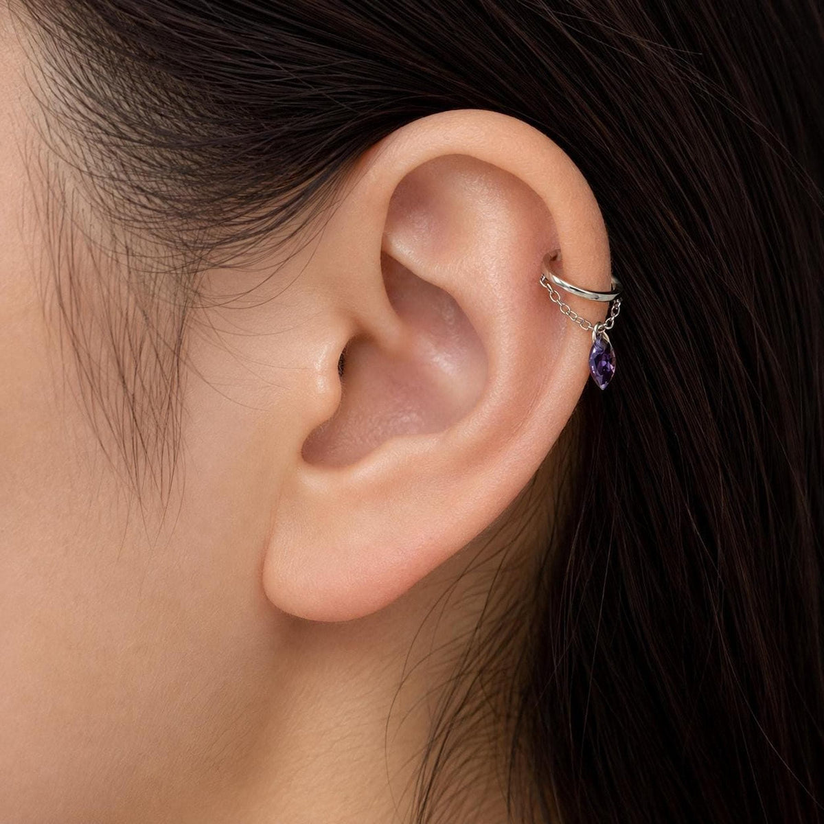 A silver ear cuff with a dangling purple marquise gem and chain worn on the cartilage of a human ear.